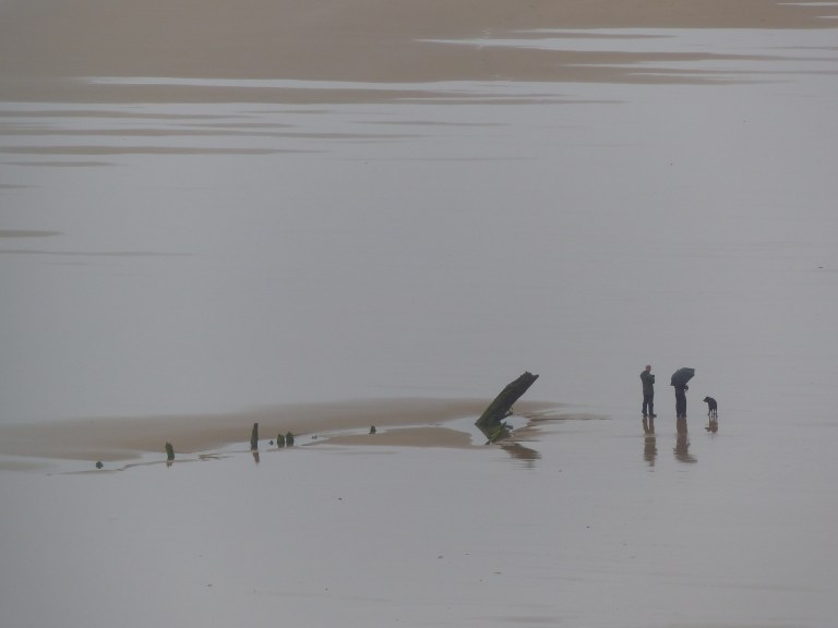 Two people with dog and umbrella by the wreck of the Helvetia on Rhossili Beach.
