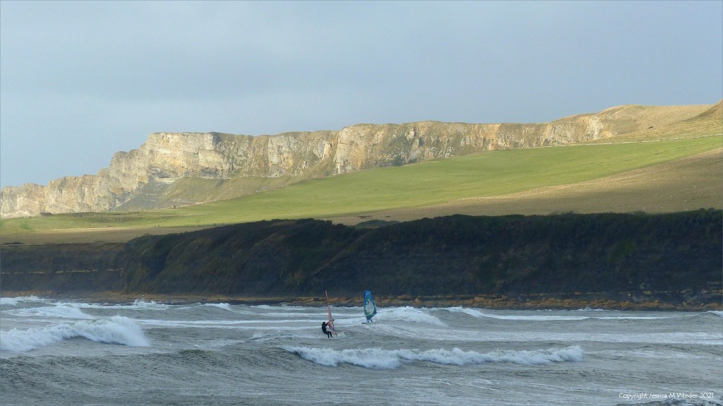 Wind surfers in the stormy waves at Kimmeridge Bay in Dorset 31 October 2021 with sunlit cliffs in the background
