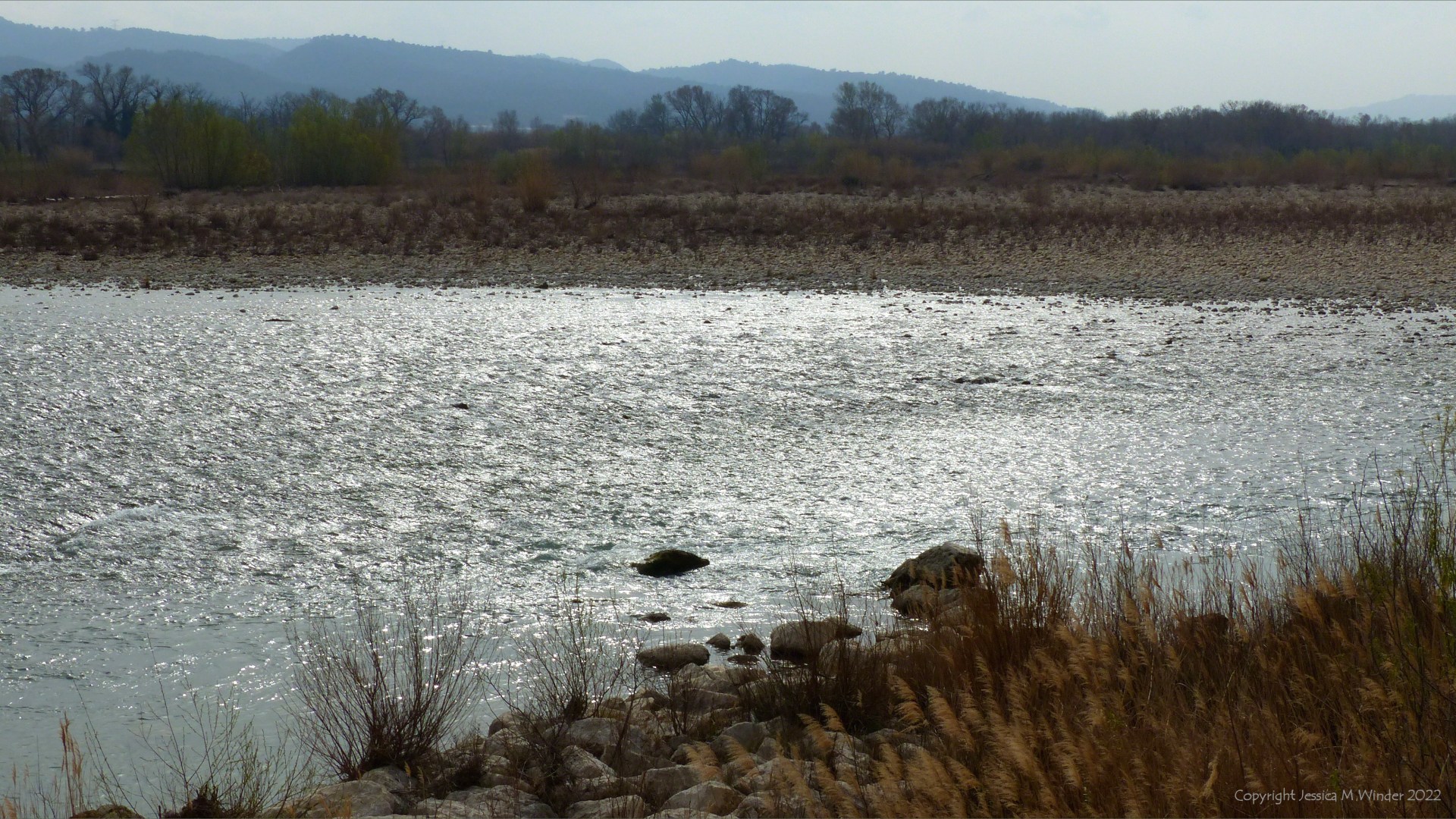 Landscape with riverbank vegetated pebbles, trees, and hills in the background on a dull early spring day